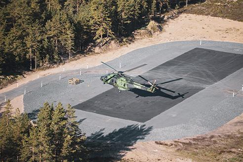 NH-90 helikopteri laskeutumassa tankkauspaikalle, joka on asfaltoitu, laaja kenttä suoja-alueineen. Takana metsää