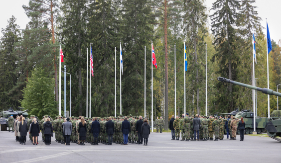 Henkilöstöä seisomassa selät kameraan päin. Tanskan, Yhdysvaltojen, Suomen, Norjan ja Ruotsin liput liehuvat henkilöstön takana. Oikealla taustalla myös Suomen ja Naton liput.