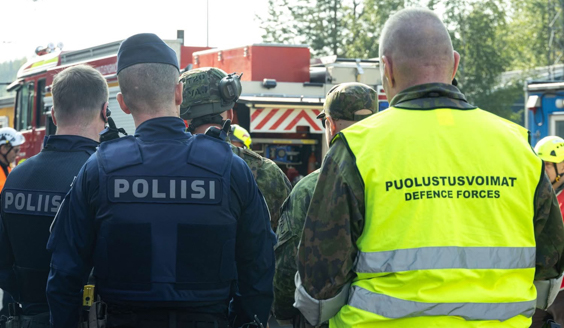 Poliisi ja Puolustusvoimien henkilökuntaan kuuluva seisovat selkä kameraan päin niin, että molempien huomioliiveissä lukevat