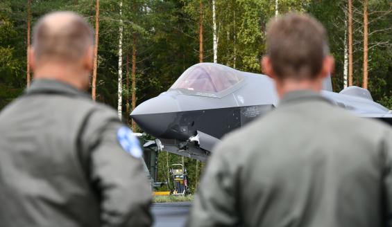 Kuvan etualalla selin kaksi miestä, Suomen ja Norjan ilmavoimien komentajat, jotka katsovat taustalla terävänä näkyvää norjalaista F-35-hävittäjää. Hävittäjän taustalla näkyy suomalaista sekametsää.