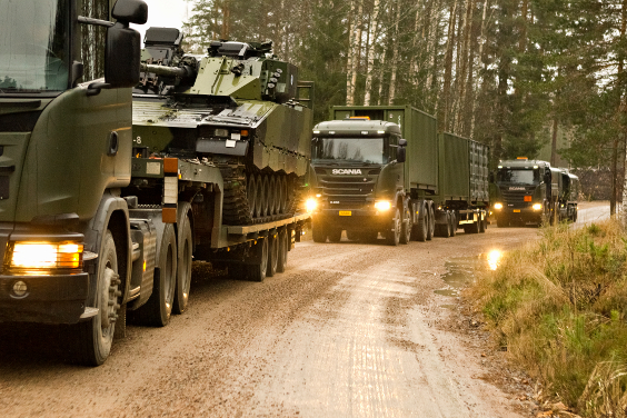 Gröna lastbilar med släp i en konvoj. Deras last består av en pansarskyttebandvagn och containrar. Foto Försvarsmakten.