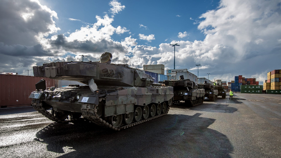 Four Leopard main battle tanks standing in line at the harbour.  The sun is shining, white clouds in the blue sky, containers of different colours in the background. Photo by Finnish Defence Forces.