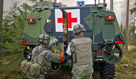 Two soldiers wearing combat uniforms and helmets are closing an armoured medical vehicle’s back door which has a red cross on it. Photo by Finnish Defence Forces.