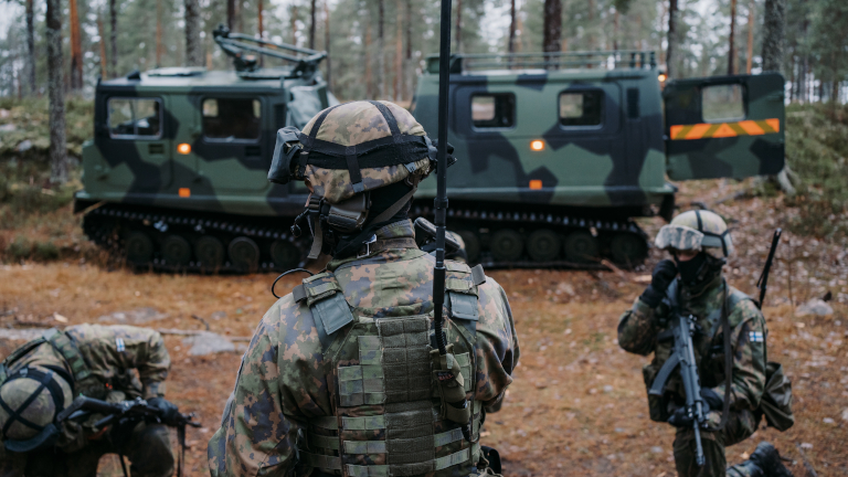 Kenttäradiota selässään kantava sotilas ja rynnäkkökivääriä kantava sotilas telakuorma-auton edessä